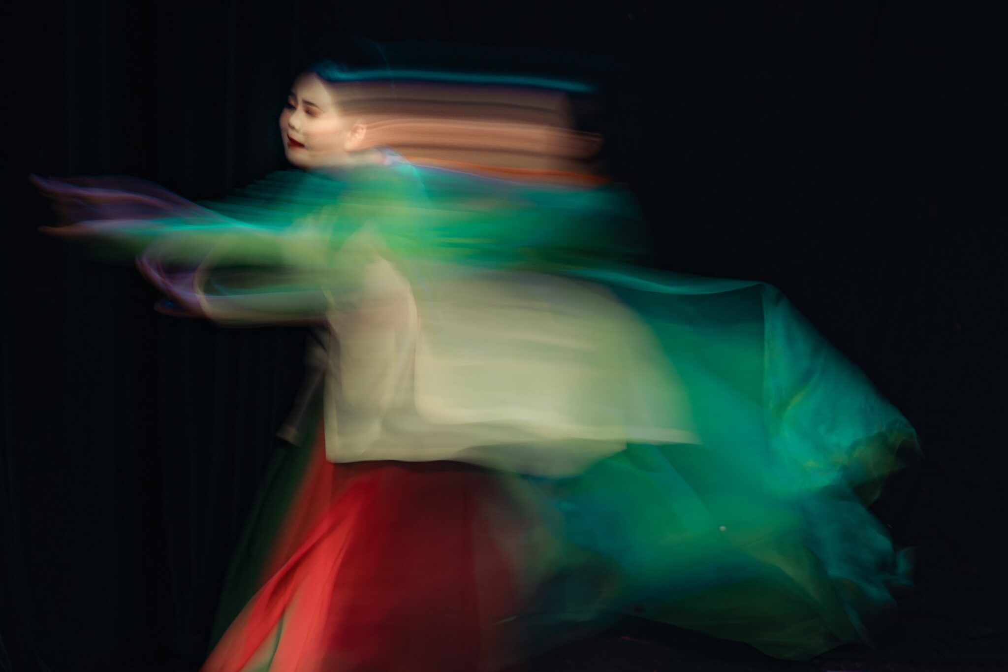 Long-exposure blur of a performer in green and red costume moving across a dark stage