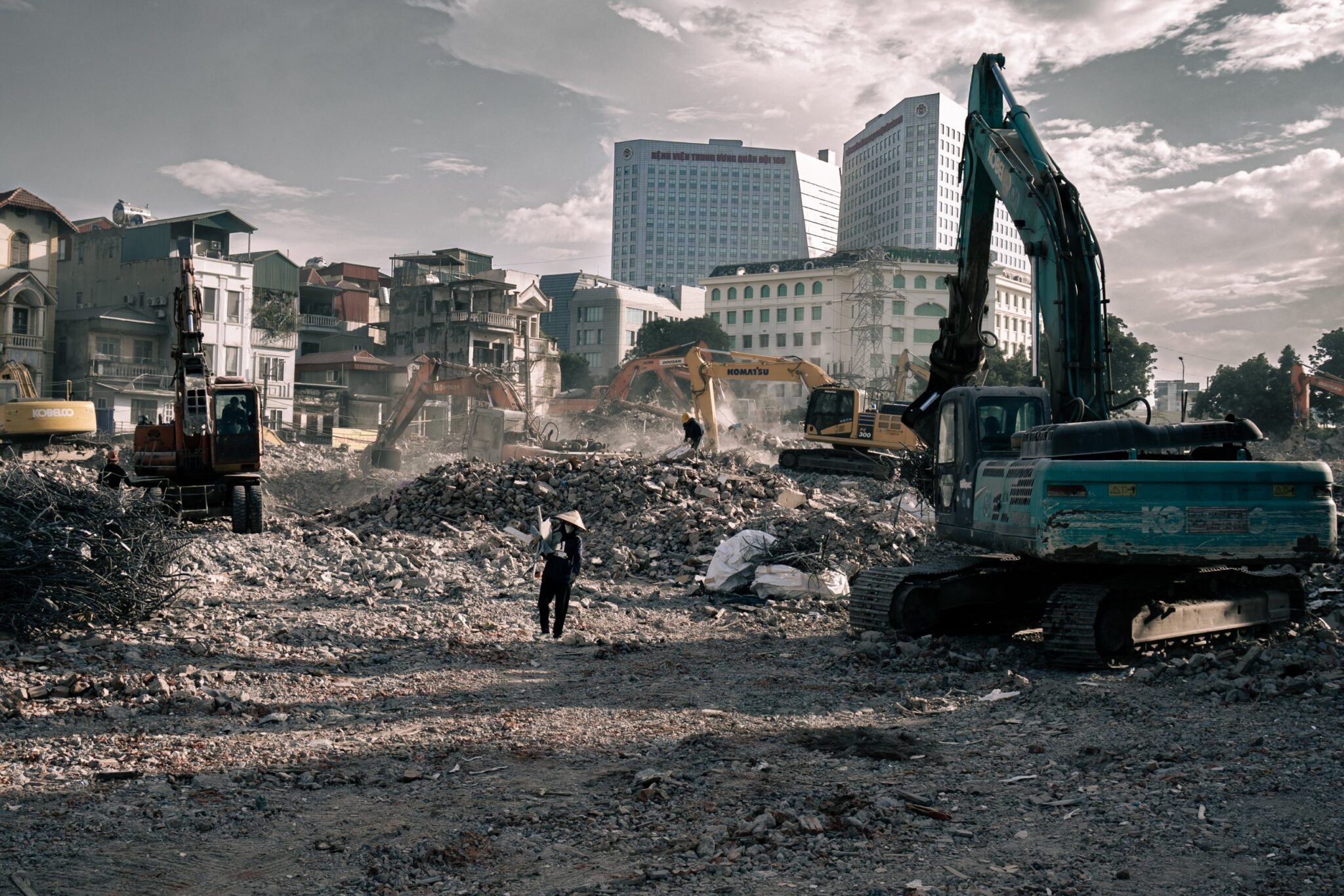 Woman in a conical hat crossing a cleared demolition site among excavators