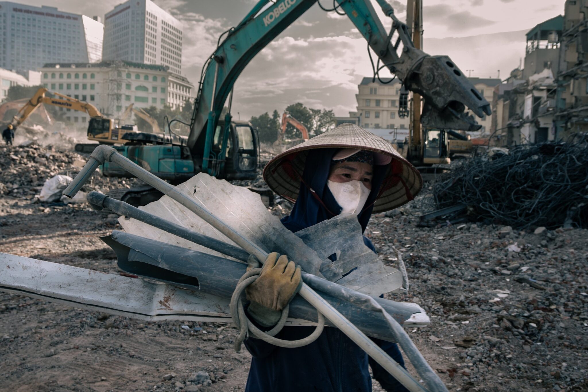 Woman in a mask and conical hat carrying salvaged pipes and sheeting