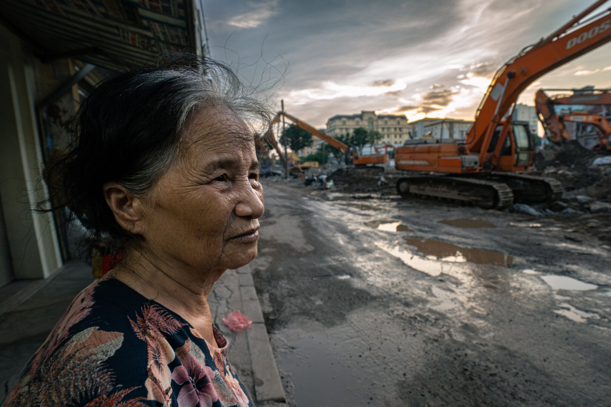 Elderly woman watching excavators clear her street at dusk