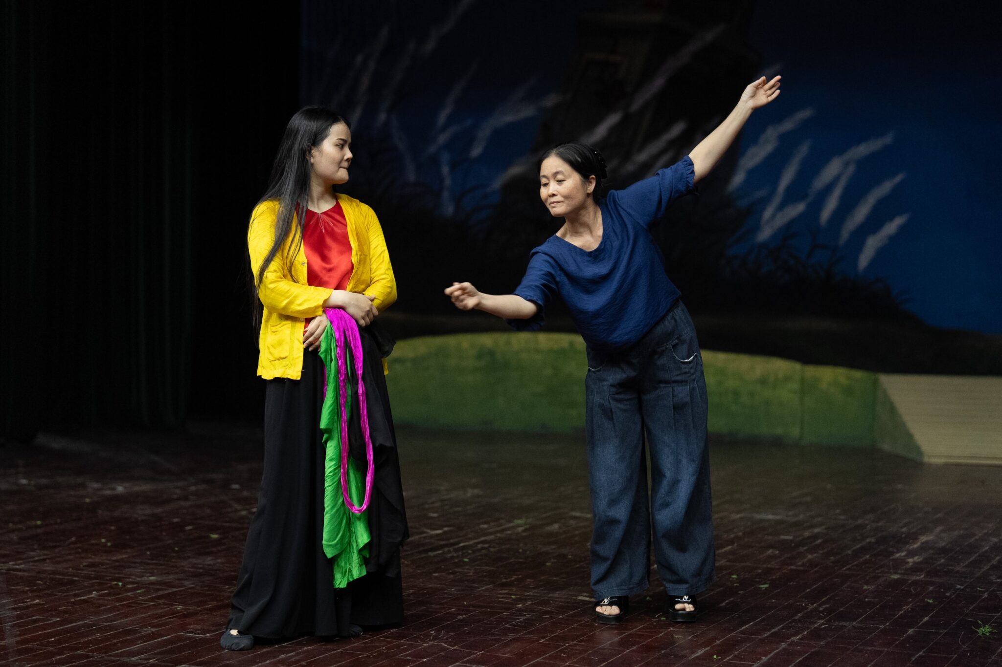 Teacher demonstrating a movement to an actress in a yellow jacket during rehearsal